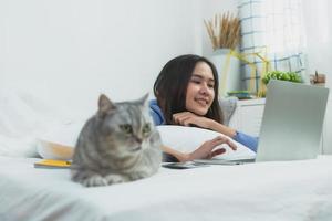 Mulher asiática trabalhando em um laptop deitada ao lado do gato na cama no quarto foto