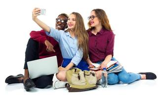 três feliz alunos com livros, computador portátil, bolsas e faz selfie foto