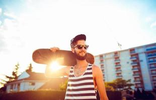 homem dentro oculos de sol com uma skate em uma rua dentro a cidade às pôr do sol luz foto