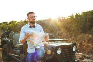 jovem à moda homem com óculos e arco gravata Vejo a mapa perto a antiquado suv foto