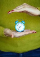 pequeno azul alarme relógio dentro a mãos do pensativo jovem mulher foto