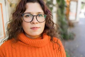 retrato do despreocupado jovem mulher sorridente com urbano fundo. alegre latim menina vestindo Óculos dentro a cidade. feliz morena mulher com encaracolado cabelo e óculos sorridente. foto