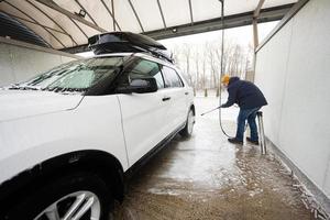 homem lavando carro suv americano de água de alta pressão com rack de teto na lavagem de autoatendimento em clima frio. foto