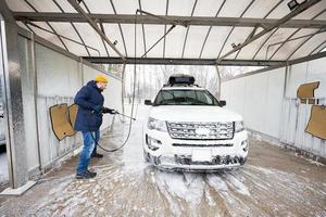 homem lavando carro suv americano de água de alta pressão com rack de teto na lavagem de autoatendimento em clima frio. foto