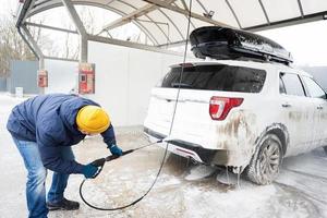 homem lavando carro suv americano de água de alta pressão com rack de teto na lavagem de autoatendimento em clima frio. foto