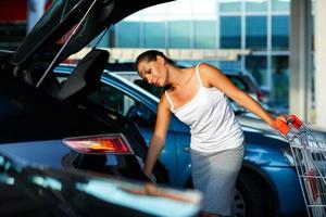 mulher turnos a compra a partir de compras carrinho dentro a tronco do uma carro em a estacionamento foto