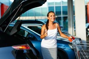 mulher em pé com compras carrinho e polegar acima perto a carro em a estacionamento foto