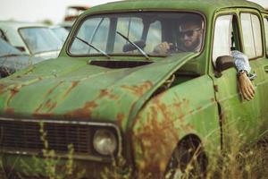 jovem à moda bonito homem, vestindo camisa e oculos de sol, dirigindo velho carro foto