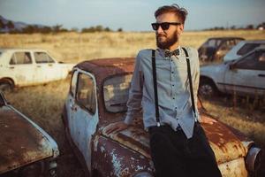 jovem bonito à moda homem, vestindo camisa e gravata-borboleta em a campo do velho carros foto