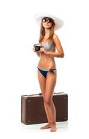 cheio comprimento retrato do uma lindo jovem mulher posando dentro uma bikini com uma Câmera dentro mãos e mala de viagem em branco foto