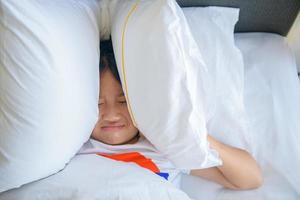 pequeno menina deitado dentro cama cobertura cabeça com travesseiro Porque também alto irritante ruído foto