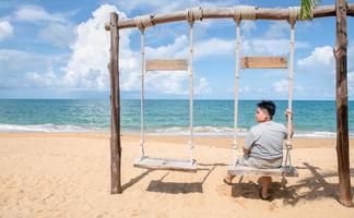 feliz turista sentar e relaxar em a de madeira balanço dentro frente do a praia, relaxar e descansar foto