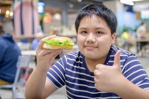 obeso Garoto apreciar para comendo carne de porco Hamburger dentro uma Comida quadra dentro uma compras Centro. foto