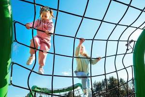 feliz ativo meninas do diferente idades escalou em uma corda rede dentro uma Parque infantil contra uma brilhante azul céu em uma ensolarado dia. foto