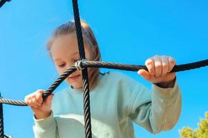 uma feliz ativo dez ano velho menina escalou uma corda rede em uma Parque infantil contra uma brilhante azul céu em uma ensolarado dia foto