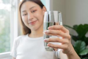 feliz lindo, ásia jovem mulher, menina bebendo, trago fresco vidro do água para hidratação do corpo, segurando transparente vidro dentro dela mão, com sede às lar. saúde Cuidado, saudável estilo de vida conceito. foto