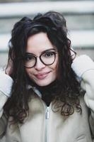 lindo jovem mulher com morena encaracolado cabelo, retrato dentro olho óculos desfrutando a Sol dentro a cidade. foto