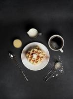 topo Visão do saboroso waffles placa, caramelo molho, café xícara, leite, colher de sobremesa, filtro em uma Preto concreto fundo foto