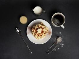 topo Visão do saboroso waffles placa, caramelo molho, café xícara, leite, colher de sobremesa, filtro em uma Preto concreto fundo foto