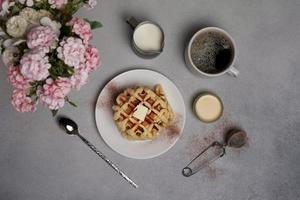 topo Visão do saboroso waffles placa, caramelo molho, café xícara, leite, colher de sobremesa, filtro, Rosa flores em uma cinzento concreto fundo foto