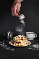 homem preparado café da manhã, delicioso waffles, caramelo molho, café xícara, leite, sobremesa colher, filtro em Preto concreto fundo foto