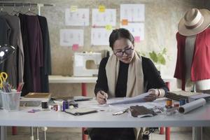 designer de moda feminina asiática de meia-idade trabalhando em um estúdio por meio de ideias desenhando esboços com fios coloridos e costurando para uma coleção de design de vestidos, alfaiate profissional empresária de sme. foto