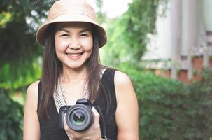 mulher asiática, usando chapéu e top preto sem mangas, de pé no jardim e segurando a câmera dslr, sorrindo alegremente. foto