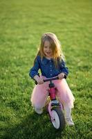 uma feliz lindo pequeno menina corre velozes e passeios dela primeiro bicicleta sem pedais em a Relva em uma caloroso verão dia. fofa bebê 3 anos velho. lado visualizar. Aprendendo para manter equilíbrio. foto