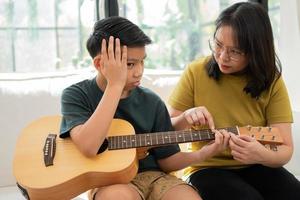 mãe asiática abraça filho, menino asiático tocando violão e mãe abraçada no sofá e se sente apreciada e encorajada. conceito de uma família feliz, aprendizado e estilo de vida divertido, ama os laços familiares foto