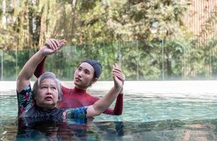 jovem treinador ajudando Senior mulher dentro aqua aeróbica e trabalhando Fora dentro a piscina. velho mulher e maduro homem fazendo aqua aeróbica exercício dentro natação piscina, idosos Esportes, e ativo estilo de vida conceito. foto