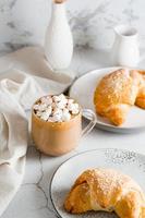 copo do café com marshmallows e croissants em pratos em a mesa. caseiro café da manhã estilo de vida. vertical Visão