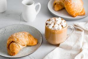 copo do café com marshmallows e croissants em pratos em a mesa. caseiro café da manhã estilo de vida foto