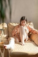 garota com uma perna engessada, sentada no sofá e desenhando em uma mesa digitalizadora. perna enfaixada e dedos dos pés após um acidente de ferimento em execução. Garota adolescente em um molde de gesso. foto