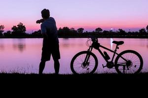 homem exercício perto bicicleta em pôr do sol e lago fundo foto