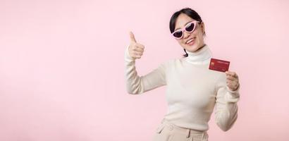 fascinante Diversão alegre jovem mulher do ásia etnia 20s anos velho com vestem oculos de sol desgasta branco camisa aguarde dentro mão crédito banco cartão isolado em avião pastel luz Rosa fundo estúdio retrato. foto