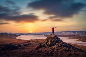 viajante homem vestem vermelho roupas e levantando braço em pé em montanha às pôr do sol dentro lago Baikal, Sibéria, Rússia. foto