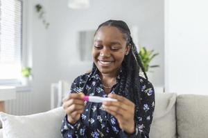 jovem mulher olhando às gravidez teste dentro felicidade. finalmente grávida. atraente Preto mulheres olhando às gravidez teste e sorridente enquanto sentado em a sofá às casa foto