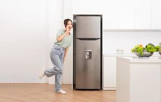 ásia mulher em pé Próximo para a geladeira dentro a cozinha foto