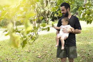 Papai e bebê dentro a parque. bebê menina toques a verde folhas do a árvores foto