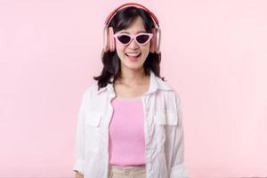 feliz jovem ásia mulher modelo com à moda na moda Sol óculos apreciar ouvindo música de fone de ouvido audio e dançando isolado em Rosa estúdio fundo. tecnologia, menina moda, acessório conceito. foto