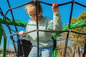 uma feliz ativo dez ano velho menina escalou uma corda rede em uma Parque infantil contra uma brilhante azul céu em uma ensolarado dia foto