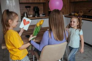 filhas felicitar seus mãe em mãe dia, uma cartão com uma coração, flores e uma balão às casa dentro a cozinha. crianças surpresa seus mãe para a feriado. foto