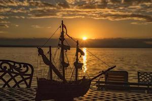 uma silhueta do uma cadeira e navio brinquedo em uma mesa às nascer do sol às a mar de praia foto