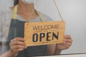 proprietário o negócio varejo, ásia jovem mulher mão dentro girando placa borda para aberto café comprar, vestindo face mascarar proteger para pandemia do coronavírus, reabrir loja depois de fechar bloqueio quarentena dentro COVID-19. foto