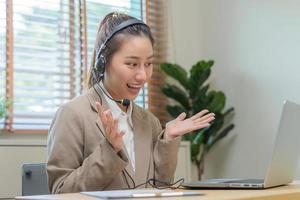 operador ásia jovem mulher vestindo fone de ouvido, fones de ouvido e Falando em vídeo ligar conferência com cliente, colegas Apoio, suporte telefone, trabalhos em computador portátil informática do ajuda, consultar serviço foto