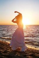 lindo mulher posando em a de praia às pôr do sol dentro uma branco vestir foto