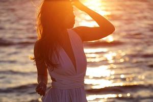 lindo mulher posando em a de praia às pôr do sol dentro uma branco vestir foto