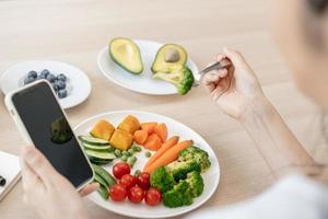 fazer dieta, ásia jovem mulher comendo, segurando garfo às brócolis, dieta plano nutrição com fresco legumes salada apreciar refeição enquanto usando Smartphone. nutricionista do saudável, nutrição do peso perda foto