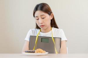 dieta, atraente ásia jovem mulher, menina contido para comer rosquinha, padaria e frito frango, velozes Comida para perder, perda peso, abraçando peso balanças em mesa às lar. paixão, tentação quando com fome. foto