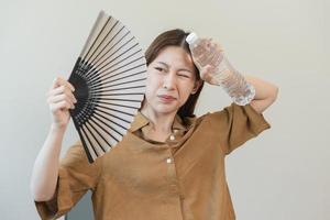 verão calor AVC, quente clima, cansado ásia jovem mulher suado e sedento, refrescante com mão dentro sopro, onda ventilador para ventilação, segurando frio água garrafa toque dela corpo quando temperatura alto. foto